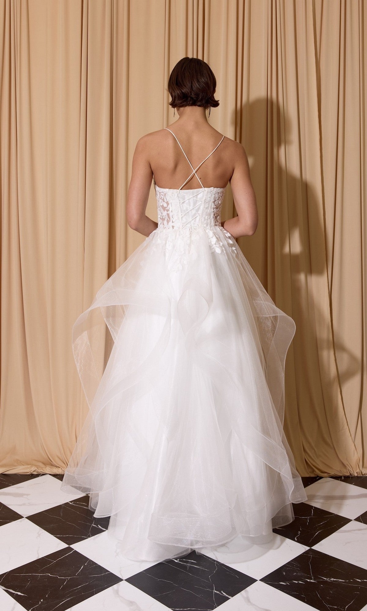 A woman wears the Eureka EK167 long prom dress, featuring white glitter tulle, a floral lace embroidered corset, and crisscross straps, while standing on a black and white checkered floor before beige curtains.